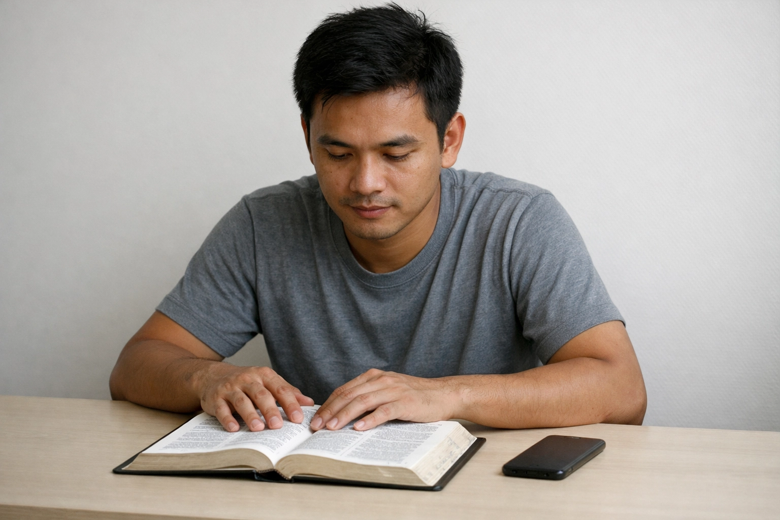 A young Southeast Asian adult sitting at a simple desk with an open Bible and a smartphone placed face down in calm, neutral daylight.